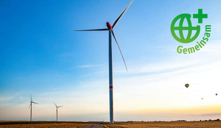 Eine große Windkraftanlage steht in einer offenen Landschaft unter einem klaren blauen Himmel. Im Hintergrund sind weitere Windräder und Heißluftballons zu sehen. In der oberen rechten Ecke befindet sich ein grünes Logo mit dem Wort „Gemeinsam“, das für nachhaltige Energie und Zusammenarbeit steht.