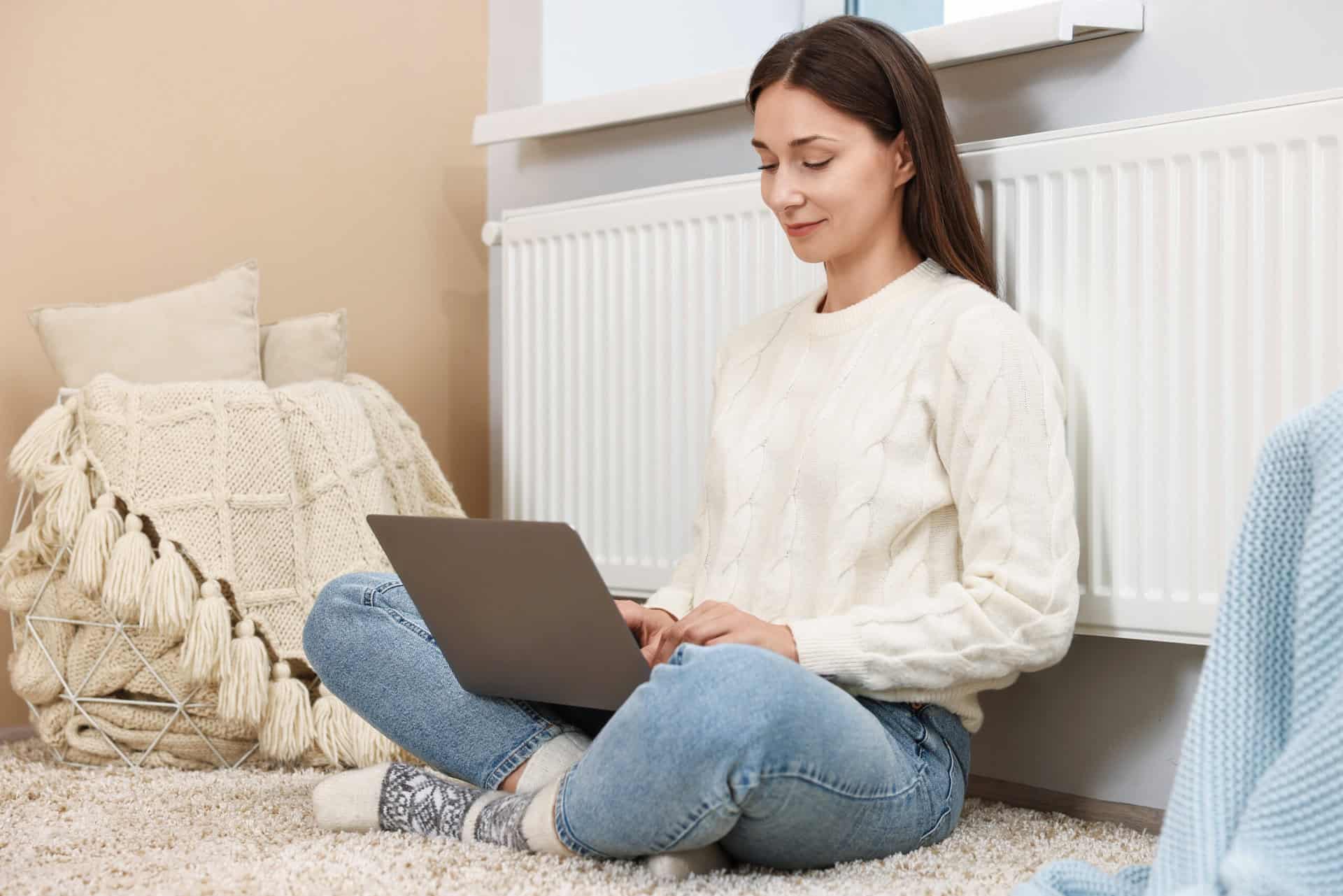 Glückliche Frau sitzt zu Hause in der Nähe eines Heizkörpers und nutzt ihren Laptop, während sie die Wärme genießt.
