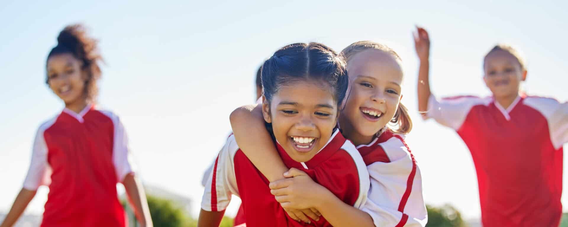 Kinder in roten Fußballtrikots lachen und spielen gemeinsam auf einem Sportplatz – Symbol für Teamgeist, Nachwuchsförderung und regionales Engagement.