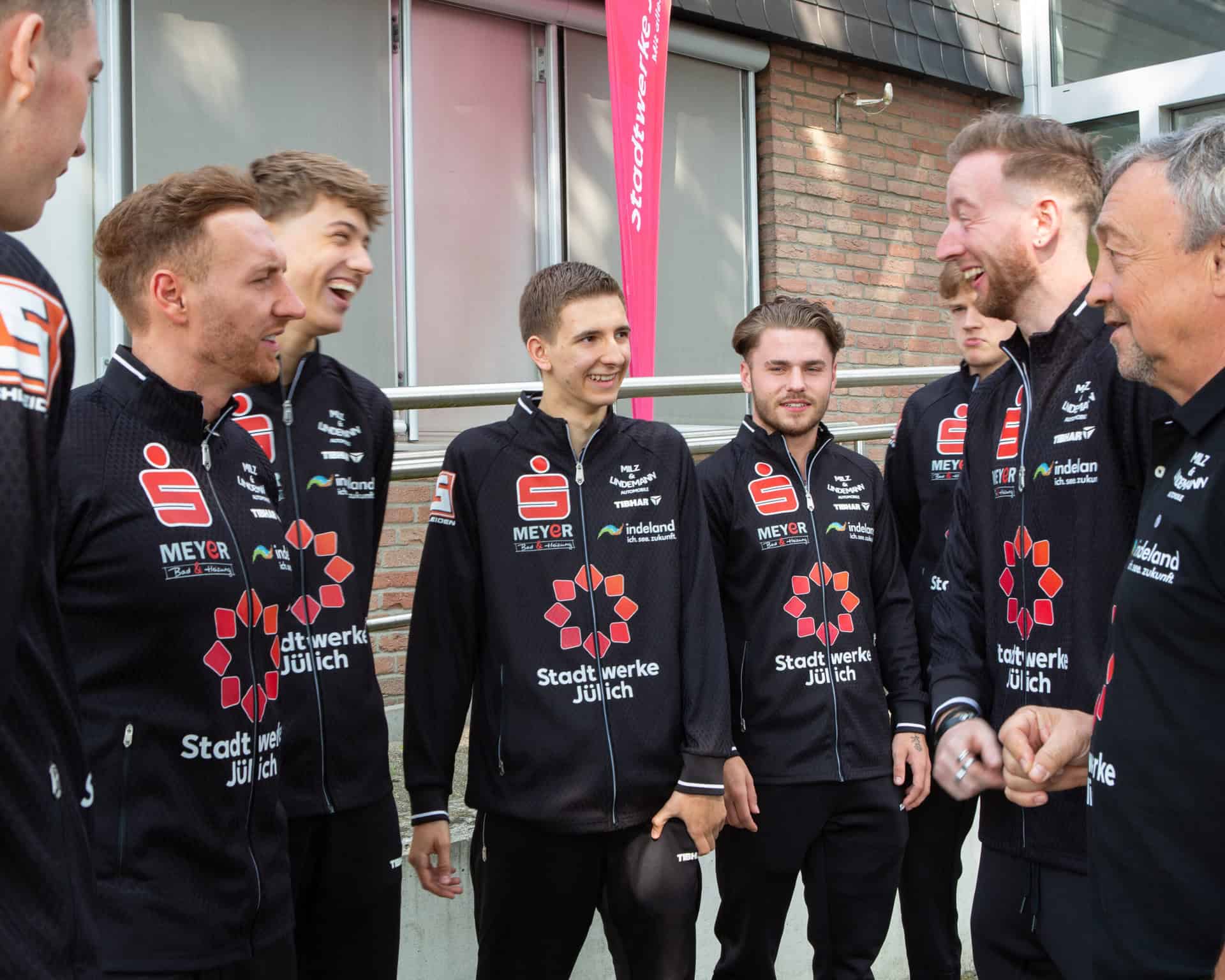 Die Tischtennismannschaft des TTC Indeland Jülich im Stadtwerke-Trikot – lächelnd im Gespräch vor einer Sporthalle.