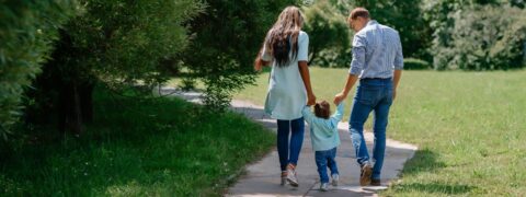 Junge Familie spaziert Hand in Hand auf einem Weg im Grünen – Symbol für Versorgung und Gemeinschaft