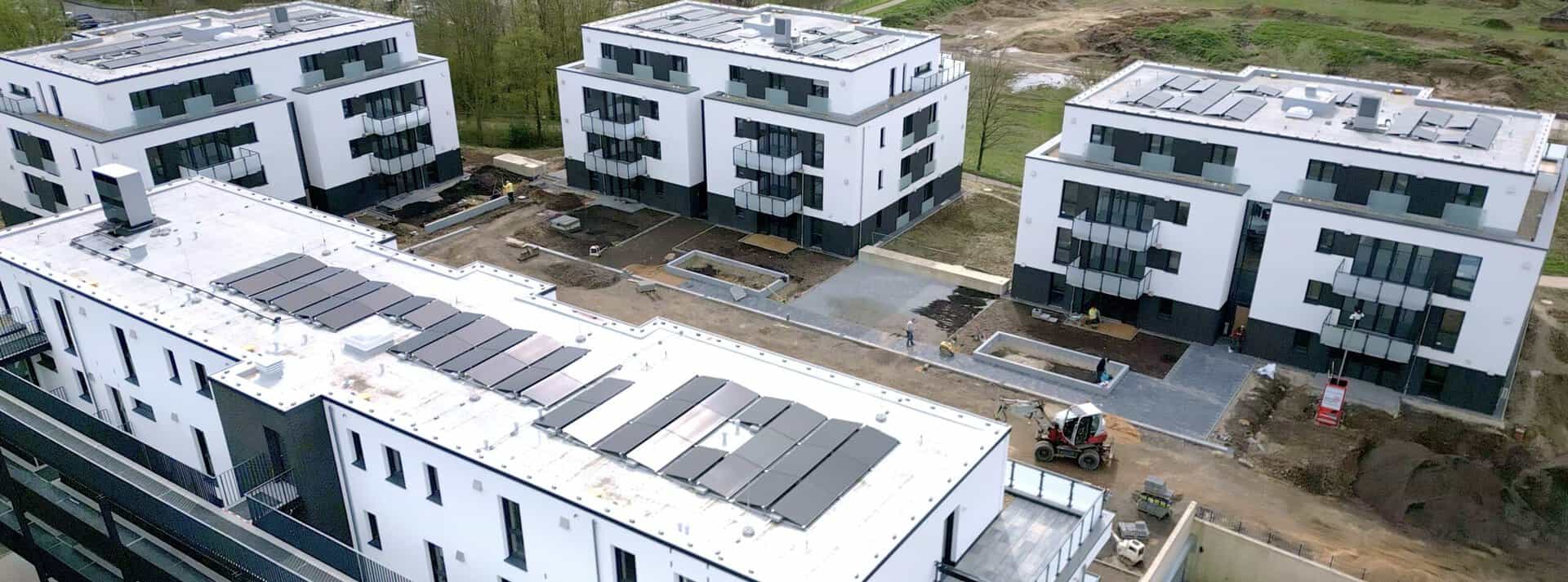 Moderne, weiße Wohngebäude mit großen Fenstern und Balkonen befinden sich in einer Neubausiedlung. Auf den Flachdächern sind Solarpaneele installiert. Der Außenbereich ist noch in der Fertigstellung, mit Bauarbeitern und Baumaschinen vor Ort.