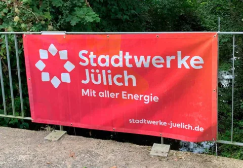 Rotes Spannband mit Logo und Claim der Stadtwerke Jülich, befestigt an einem Geländer im Grünen.