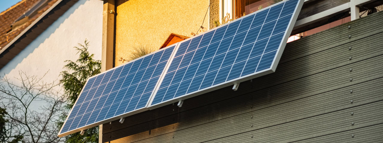 Ein Balkonkraftwerk mit zwei Solarpanelen ist an der Fassade eines Wohnhauses montiert. Die Sonne spiegelt sich in den Modulen, während ein minimalistisches Symbol für Solarenergie und nachhaltiges Wohnen in der unteren linken Ecke des Bildes zu sehen ist.