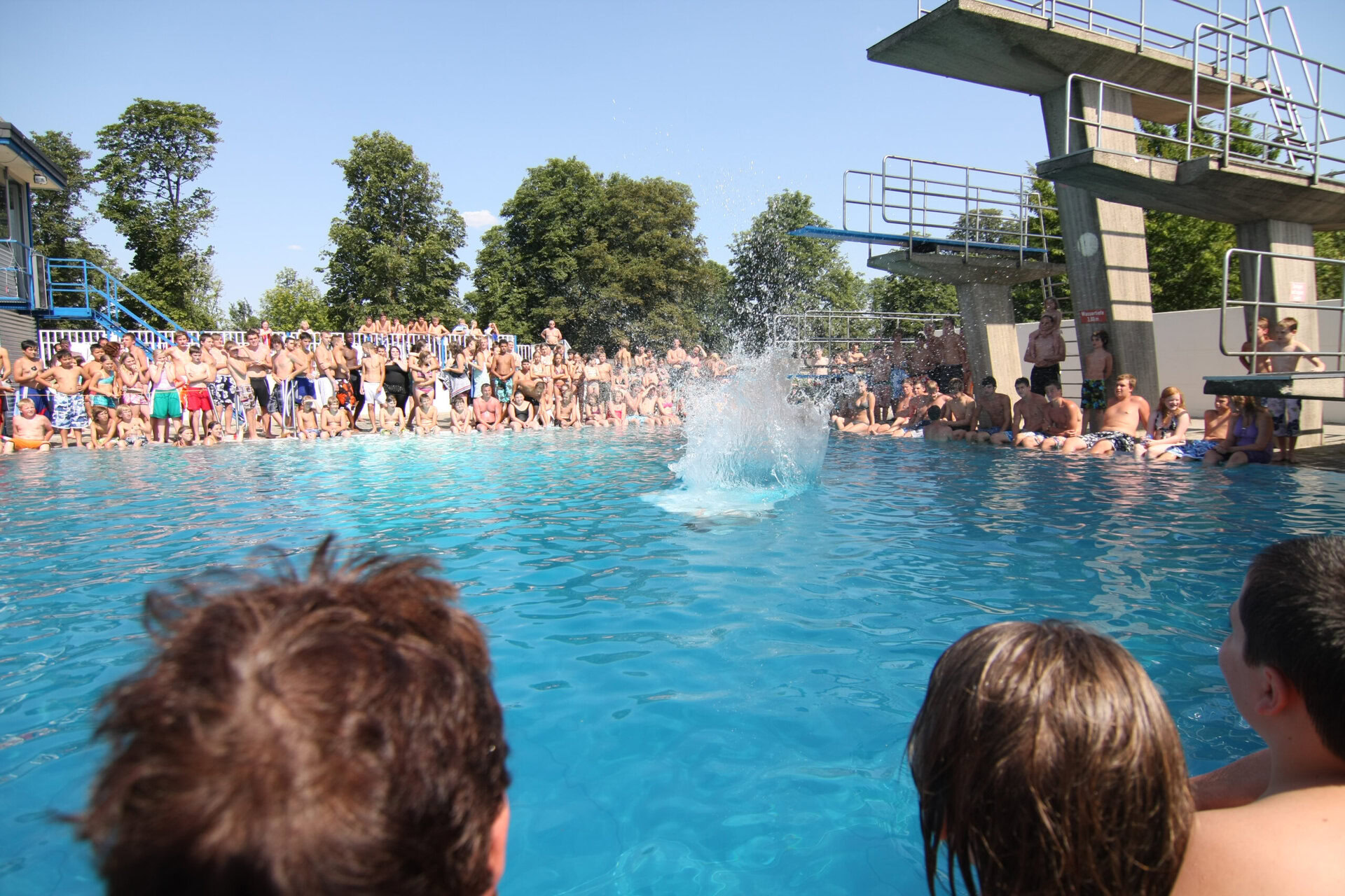 Auf dem Bild ist ein großer Sprungturm zu sehen und ein Schwimmbecken. Im Schwimmbecken ist eine Fontäne weil jemand dort vom Turm hinein gesprungen ist. Um das Schwimmbecken stehen viele Leute und Kinder die das beobachten.