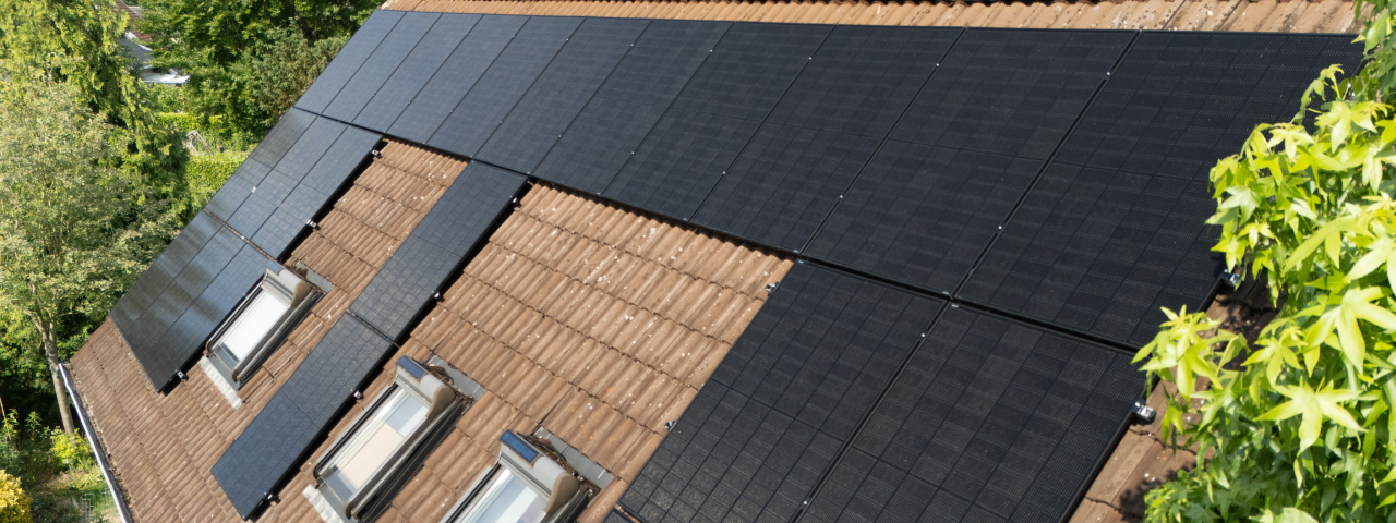 Zu sehen ist ein Dach mit braunen Ziegeln. Auf dem Dach sind Solarmodule zu sehen, die sehr neuwertig aussehen. Im Vordergrund und im Hintergrund sieht man grüne Bäume.