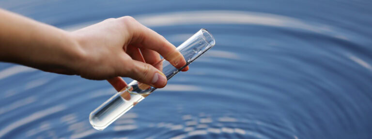 Auf dem Bild ist Wasser zu sehen und eine Hand, die mit einem Reagenzglas eine Probe entnimmt.