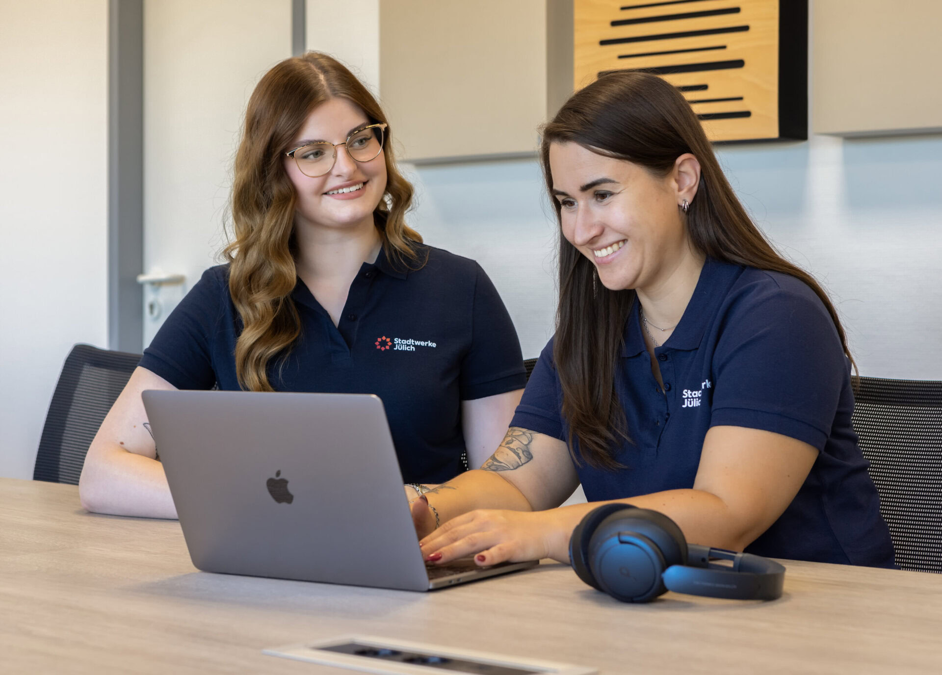 Auf dem Bild sind zwei Frauen zu sehen, die an einem Tisch sitzen und gemeinsam an einem Laptop arbeiten. Beide lachen dabei.